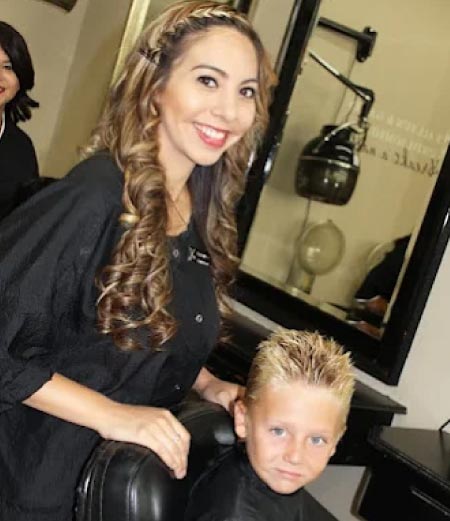 A professional female cosmetologist smiling while providing a haircut to a young boy, demonstrating the practical family-friendly services taught during the 1,000-hour cosmetology course.