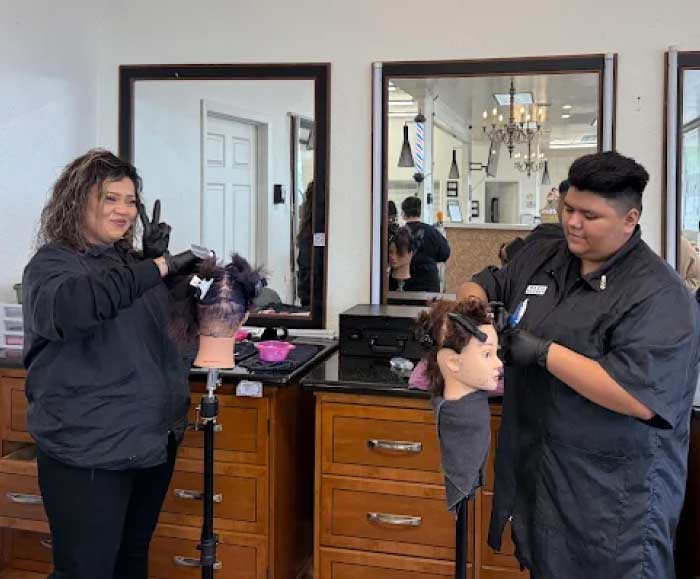 Two cosmetology students at Lawrence & Company College of Cosmetology practicing hair styling techniques on mannequin heads in a professional student salon. The student on the left wears black gloves and makes a peace sign while sectioning hair, while the student on the right focuses on a detailed cutting or styling maneuver. This hands-on training is a core part of the immersive 1000-hour program designed to prepare students for a thriving career in the beauty industry.
