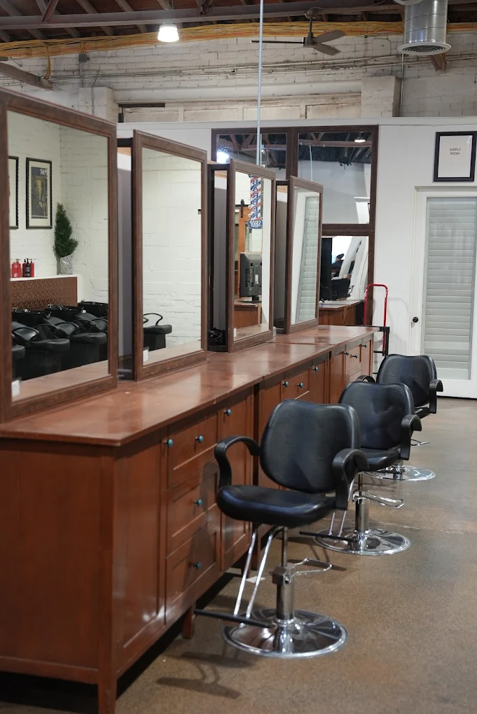 A professional barbering student salon featuring rows of wooden vanity stations with large rectangular mirrors and black hydraulic styling chairs. The background shows professional hair washing stations and an open industrial-style ceiling.