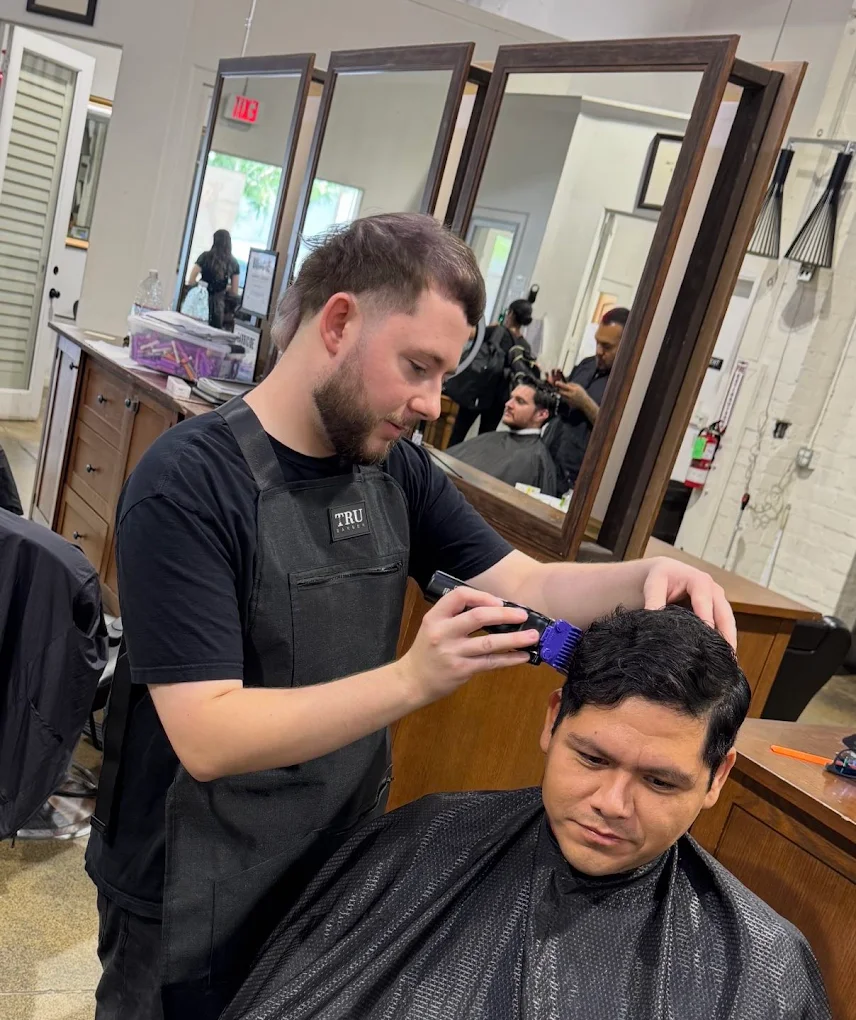 A focused barbering student wearing a black apron performing a precision haircut with electric clippers on a client. The background shows a clean, modern student salon environment with large mirrors reflecting the instructional workspace.