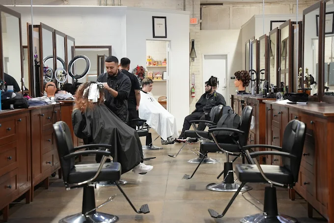 A professional barbering student salon featuring rows of wooden vanity stations with large rectangular mirrors and black hydraulic styling chairs. The background shows professional hair washing stations and an open industrial-style ceiling.